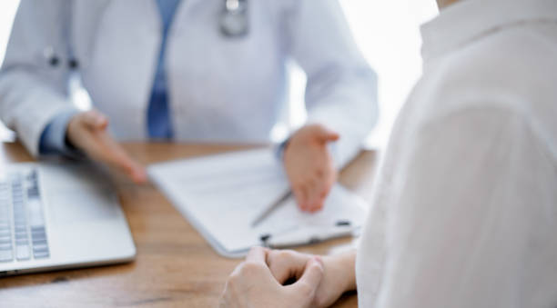 istock Doctor and patient sitting and discussing something at wooden table while using a laptop computer. Focus is on patient's hands. Medicine concept 1492399386