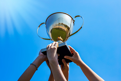 Hands holding a golden cup trophy against a blue sky with sunbeams, victory success scholl team concept