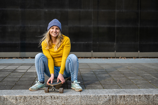 Young female teenager dressed in yellow jersey and jeans, sitting on a skateboard, looks at the camera and smiles, generation z lifestyle