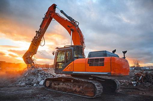 Excavator with concrete crusher on rigging and pile of reinforced cement for material recycling at demolition site at sunset