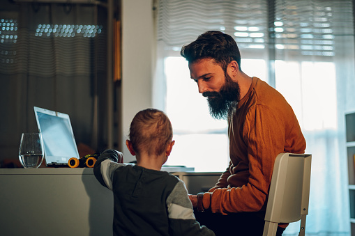 A father is getting distracted by his toddler during his remote job time at the home office. A son is bothering his freelancer father and distraught him while standing at the home office.