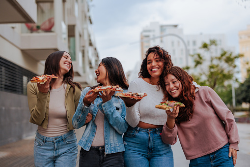 Four young diverse women in 20s bonding while eating pizza and walking down the street.