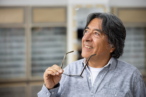 Happy Latin American senior man holding a pair of glasses at the ophthalmologist store and smiling