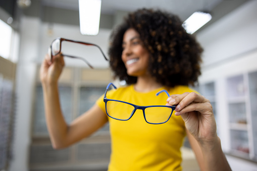 Happy African American woman trying on two pairs of glasses at the opticians shop and smiling