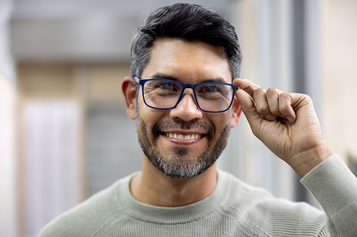 Portrait of a happy Latin American man trying on glasses at the opticians shop and smiling at the camera