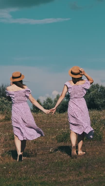 Vertical, Young Twins Sisters in Summer Dresses Walks Green Field Holding Hands