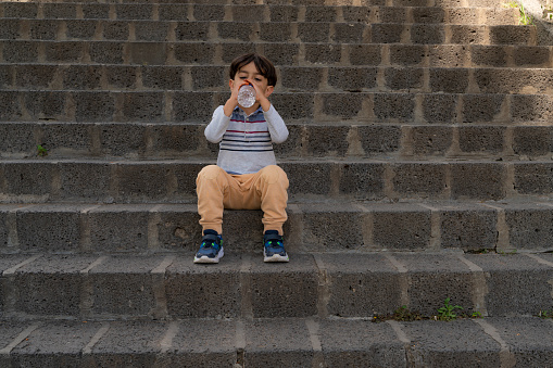 boy sitting on stone stair steps. He is drinking water. Shot with a full-frame camera in daylight.
