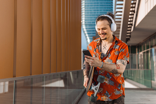 Portrait of a smiling Latin American man text messaging on his smart phone and carrying his skateboard while walking