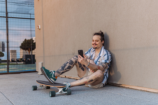 Portrait of a young tattooed Latin American man sitting on the ground with a foot on his skateboard and looking at the mobile phone