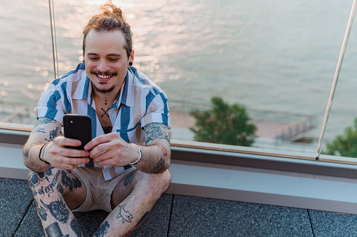 Portrait of a Latin American man with a tattooed body spending a day by the river. He is sitting comfortably on the ground and surfing the net on his smart phone.