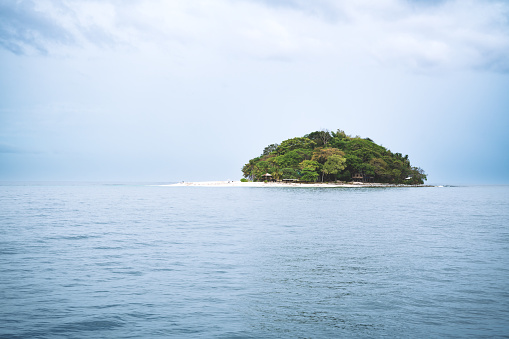 Small remote white beach island with lush green trees in light blue ocean and dramatic sky, Palawan, Philippines