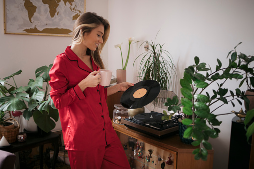 Caucasian female in nightwear holding a vintage vinyl record and drinking coffee at home in morning. Woman in pajamas looking at LP record and having morning coffee indoors.