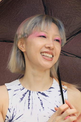 A Taiwanese model laughing during a pose under her umbrella. She is wearing short, straight multi colored hair, a blue and white pattern sleeveless dress and makeup.