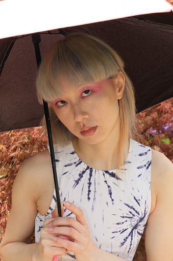 A closeup of a Taiwanese woman. She is wearing short, straight, multi colored hair, an umbrella, makeup, and a blue and white pattern sleeveless dress.
