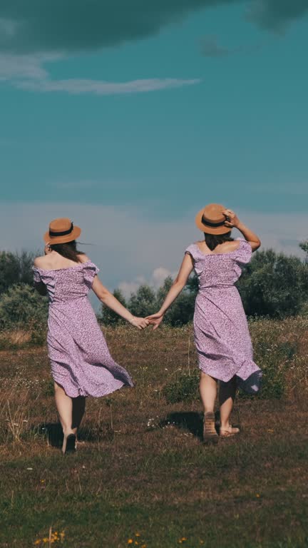 Vertical, Young Twins Sisters in Summer Dresses Walks Green Field Holding Hands