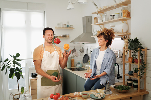 Smiling young man juggling bell peppers while making pizza with his wife in a domestic kitchen