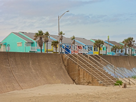 A colorful row of houses behind the famous Galveston sea wall protecting the island and city from the devastating effects of hurricane storm surge. The retaining wall rises above the beach sand that has recently been washed along the base of the wall. Periodic stairwells provide access to the beach. With condominium in the background.