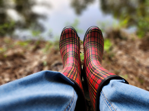Woman sitting by pond wearing plaid rubber boots.