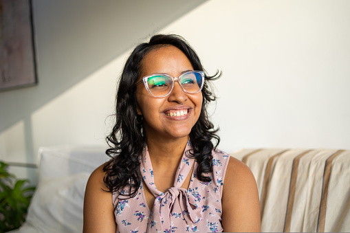 Portrait of young Hispanic woman with a glasses in the apartment with lot of sunlight looking away with a big smile. High quality photo