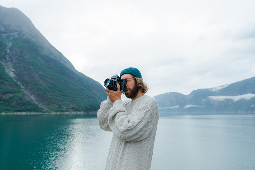 Man  in white sweater photographing Norwegian scenic landscape with film camera