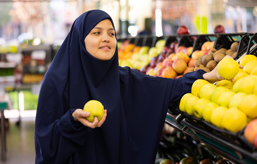 Young woman in paranja shopping and choosing local juicy apples in hypermarket
