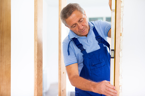 Man builder in blue overalls measures deviations from a given vertical wall using a building level