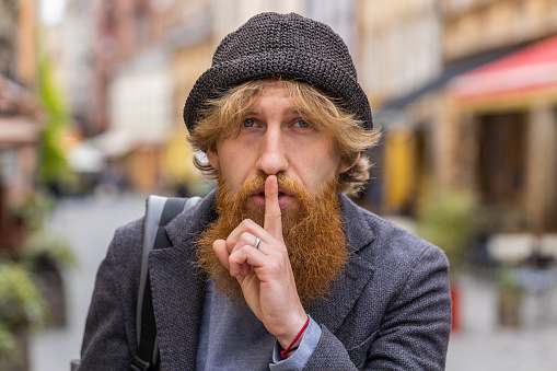 Shh be quiet please. Young bearded man presses index finger to lips makes silence hush gesture sign do not tells gossip secret. Redhead guy walking in urban city street outdoors. Town lifestyles