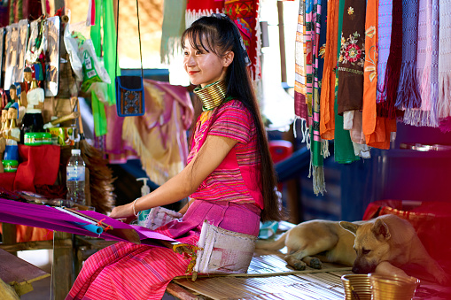 A portrait of a woman from a long-necked tribe, known for the metal rings around their necks, weaving a colorful blanket. Celebrates cultural diversity and tradition. Chiang Rai, Thailand - 09.02.2022