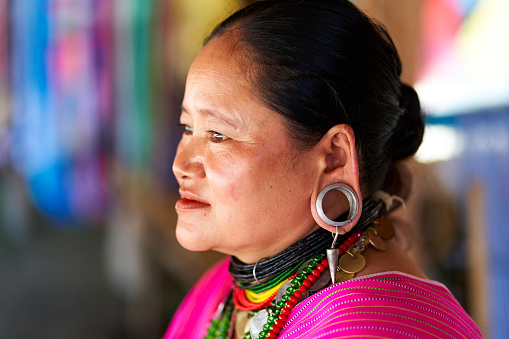 Thai woman wearing a bright fuchsia blouse, adorned with intricate jewelry around her neck. Her large ear tunnels, embellished with decorative accents, add to her unique and striking appearance. Chiang Rai, Thailand - 09.02.2022