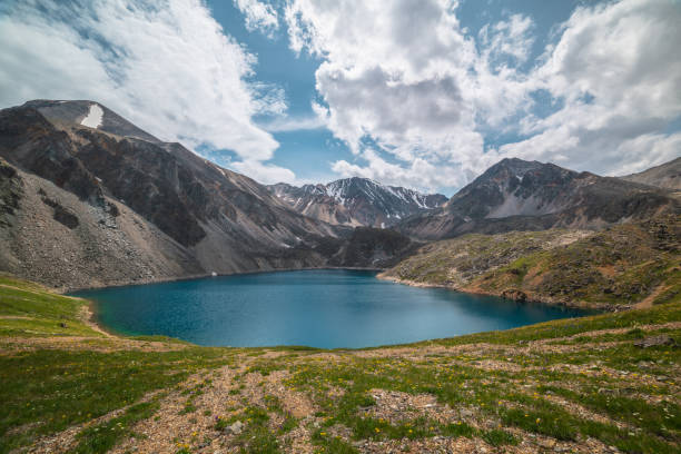 Deep mountain lake of phantom blue color among sharp rocks and high mountains in changeable weather. Wonderful dramatic view from sunlit grassy hill to deep blue alpine lake and rocks under cloudy sky
