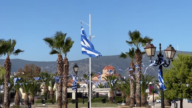 Greek flags flutter in the wind on flagpoles and streamers among palm trees against the backdrop of mountains