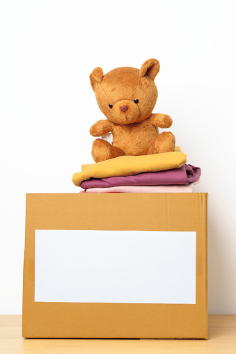 Kid Donation, Charity, Volunteer, Giving and Delivery Concept. Hand donate Bear doll and Clothes into cardboard box at home for support and help poor, refugee and homeless people. Copy space for text