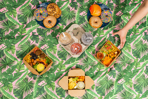 Overhead shot of a hand reaching for vegan food varieties, desserts and smoothies on a picnic blanket.