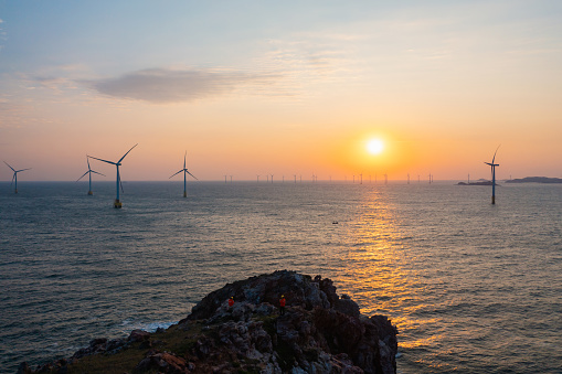 Offshore wind power plant and two electrical engineers on a rocky hill at sunrise
