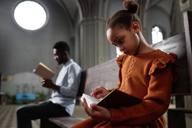 Little girl reading Bible in church African American little girl reading Bible while sitting on bench in church with man in background black gospel church stock pictures, royalty-free photos & images