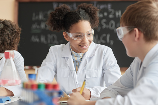 Portrait of smiling black schoolgirl wearing lab coat in science class and talking to friend.