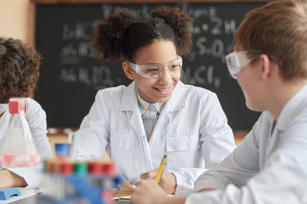 Smiling black schoolgirl wearing lab coat in science class talking to friend. Portrait of smiling black schoolgirl wearing lab coat in science class and talking to friend. school science project stock pictures, royalty-free photos & images