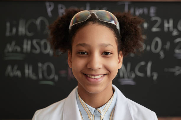 menina negra sorridente usando jaleco na aula de ciências na escola olhando para a câmera - skill child mathematics physics - fotografias e filmes do acervo