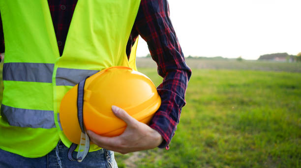 istock Construction worker holding his yellow helmet, hard hat and construction plan wearing fluorescent waistcoat on the empty field. Occupational Safety and Health OSH 1492538340