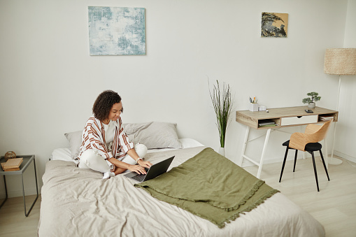 High angle portrait of black young woman using laptop on bed and smiling in cozy home interior, copy space