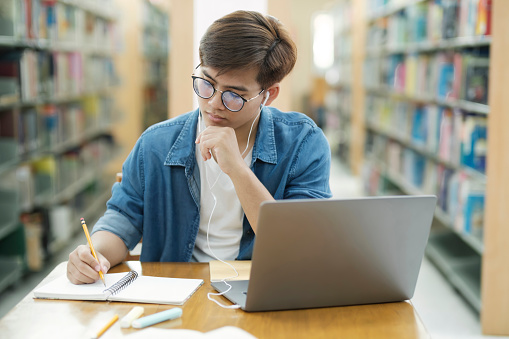 Young male college student wearing eyeglasses and in casual cloths sitting at desk studying, reading book, thinking hard, and writing down notes using laptop wearing headphones at library for research or school project. E-Learning concept.