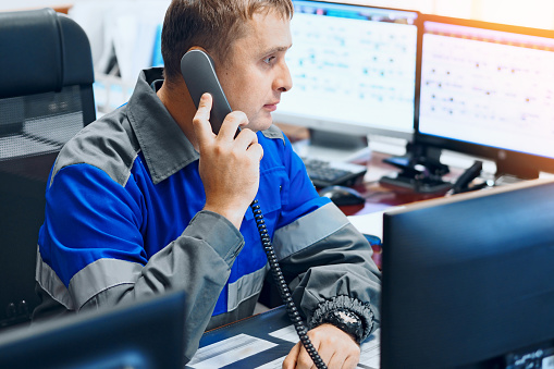 A dispatcher at an industrial enterprise sits in an office surrounded by monitors and talks on a wired telephone. Authentic workflow in the decision center of a gas company. Work with clients on gasification issues.