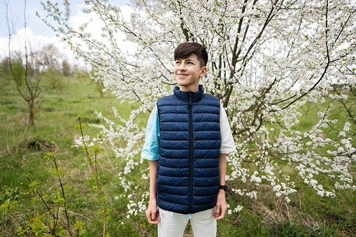 Teen boy wear sleeveless vest against white blloming tree in spring.
