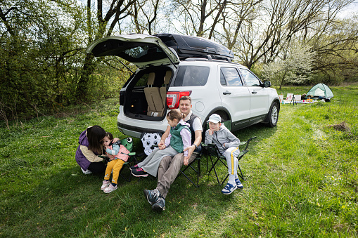 Family with kids sit on chair against car open trunk on picnic.