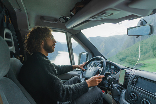 Man with curly long hair  driving camper van on scenic road in Norway that passes fjord