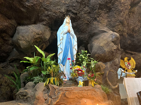 Statue of Holy Virgin Mary in Roman Catholic Church, in the cave of virgin mary, in a rock cave chapel Catholic Church with tropical flowers around in the asian