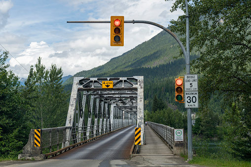 Drivers perspective of steel truss bridge across river in mountainous area of Canada with bridge deck of steel grating and signs indicating maximum height, weight and speed and traffic light on red