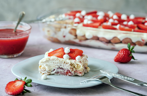 Italian traditional dessert tiramisu with strawberries. One piece of cake on a plate closeup, whole cake and strawberry juice backside, fork and knife, rose and grey concrete background