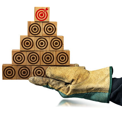 Gloved hand holding a stack of wooden blocks forming a pyramid, with many empty brown targets and one red one with an arrow reaching into the center, isolated on white background with reflections.