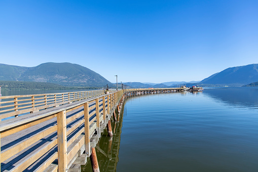 Salmon Arm, BC, Canada; August 2022; View along curved wharf and pier (Salmon Arm Wharf) with boardwalk at shore of Shuswap Lake and panoramic view of surrounding mountains against clear blue sky